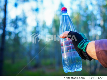 Water bottle in athlete's hand during a hike in woods. Water is used for hydration Water bottle in athlete's hand during a hike in woods. Water is used for hydration 139583273