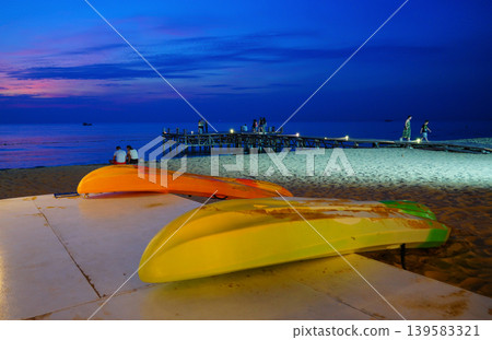 Bright kayaks sit on shore at dusk while people walk and enjoy beach scene near pier Bright kayaks sit on shore at dusk while people walk and enjoy beach scene near pier 139583321