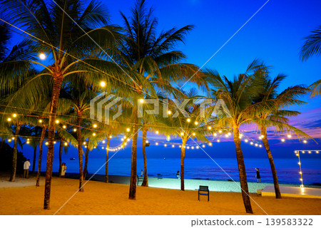 Gathering at resort beach in lights on palm trees in evening near sea. People at beach at palm trees Gathering at resort beach in lights on palm trees in evening near sea. People at beach at palm trees 139583322