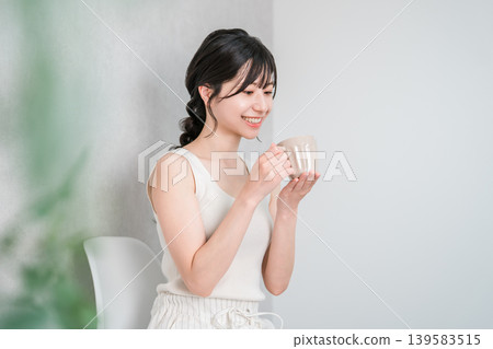 A young Asian woman in a tank top, warming herself by drinking a hot beverage from a mug, is shown in profile. A young Asian woman in a tank top, warming herself by drinking a hot beverage from a mug, is shown in profile. 139583515