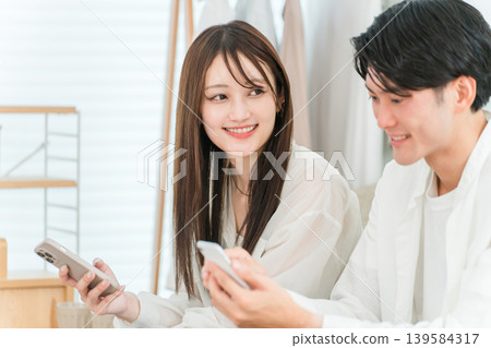 一對年輕的亞洲夫婦坐在家裡的客廳沙發上，一邊看著智慧型手機一邊聊天。 139584317