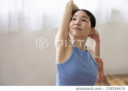 A middle-aged Japanese woman performs the pigeon pose in yoga on a mat by a bright window. A middle-aged Japanese woman performs the pigeon pose in yoga on a mat by a bright window. 139584419