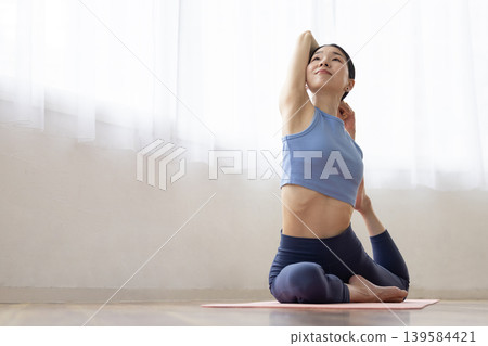 A middle-aged Japanese woman performs the pigeon pose in yoga on a mat by a bright window. 139584421