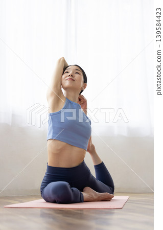 一位中年日本女性在明亮的窗邊，於瑜珈墊上練習鴿子式。 139584423