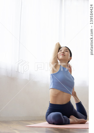 一位中年日本女性在明亮的窗邊，於瑜珈墊上練習鴿子式。 139584424