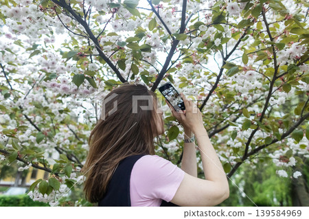 Young woman using smartphone to identify plants and take photos of blooming cherry blossom in spring park. Technology and nature photography lifestyle. Young woman using smartphone to identify plants and take photos of blooming cherry blossom in spring park. Technology and nature photography lifestyle. 139584969