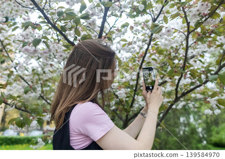 Young woman using smartphone to identify plants and take photos of blooming cherry blossom in spring park. Technology and nature photography lifestyle. Young woman using smartphone to identify plants and take photos of blooming cherry blossom in spring park. Technology and nature photography lifestyle. 139584970