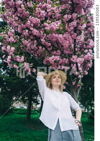 Beautiful young woman in straw hat posing near blooming pink sakura tree in spring park. Romantic girl enjoying cherry blossom outdoors. 139585063