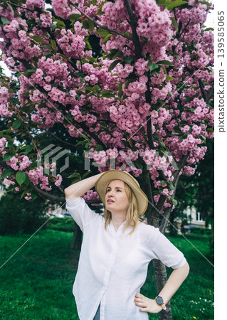 Beautiful young woman in straw hat posing near blooming pink sakura tree in spring park. Romantic girl enjoying cherry blossom outdoors. 139585065