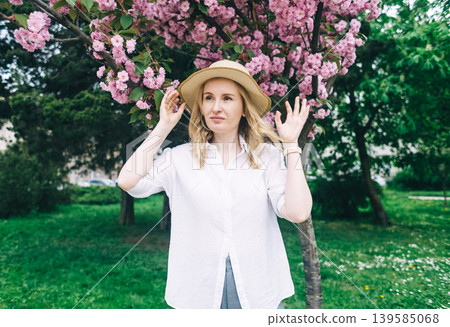 Beautiful young woman in straw hat posing near blooming pink sakura tree in spring park. Romantic girl enjoying cherry blossom outdoors. 139585068