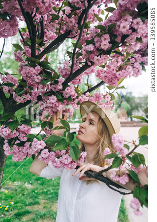 Beautiful young woman in straw hat posing near blooming pink sakura tree in spring park. Romantic girl enjoying cherry blossom outdoors. 139585085