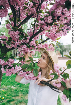 Beautiful young woman in straw hat posing near blooming pink sakura tree in spring park. Romantic girl enjoying cherry blossom outdoors. Beautiful young woman in straw hat posing near blooming pink sakura tree in spring park. Romantic girl enjoying cherry blossom outdoors. 139585086