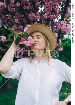Beautiful young woman in straw hat posing near blooming pink sakura tree in spring park. Romantic girl enjoying cherry blossom outdoors. 139585090