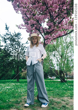 Beautiful young woman in straw hat posing near blooming pink sakura tree in spring park. Romantic girl enjoying cherry blossom outdoors. Beautiful young woman in straw hat posing near blooming pink sakura tree in spring park. Romantic girl enjoying cherry blossom outdoors. 139585108