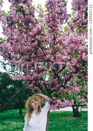 Beautiful young woman in straw hat posing near blooming pink sakura tree in spring park. Romantic girl enjoying cherry blossom outdoors. 139585111