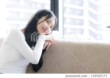 A woman lost in thought by the window; leaning back on the sofa with a smile, contemplating. This image could also evoke thoughts of buying an apartment. A woman lost in thought by the window; leaning back on the sofa with a smile, contemplating. This image could also evoke thoughts of buying an apartment. 139585171