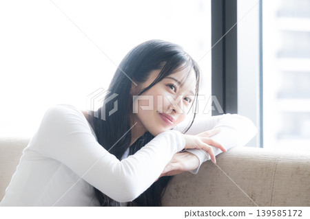 A woman lost in thought by the window; leaning back on the sofa with a smile, contemplating. This image could also evoke thoughts of buying an apartment. A woman lost in thought by the window; leaning back on the sofa with a smile, contemplating. This image could also evoke thoughts of buying an apartment. 139585172