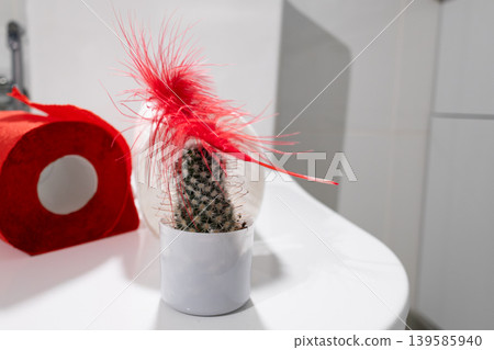 Toilet paper roll beside cactus with red feathers on bathroom counter. Playful symbol of irritation relief and hygiene in personal care. Hemorrhoid prevention concept using everyday items for comfort. Toilet paper roll beside cactus with red feathers on bathroom counter. Playful symbol of irritation relief and hygiene in personal care. Hemorrhoid prevention concept using everyday items for comfort. 139585940