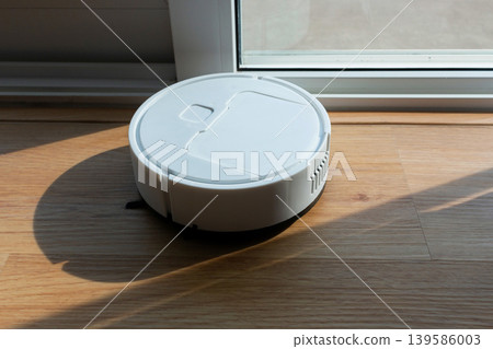 White Robot Vacuum Cleaner Working on Wooden Floor with Sunlight 139586003