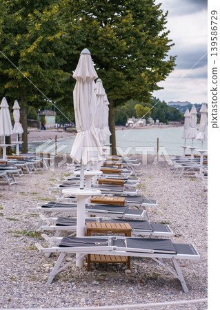 Rows of empty sunbeds and closed umbrellas on pebble beach, peaceful lakeside atmosphere with trees and soft cloudy sky in background Rows of empty sunbeds and closed umbrellas on pebble beach, peaceful lakeside atmosphere with trees and soft cloudy sky in background 139586729