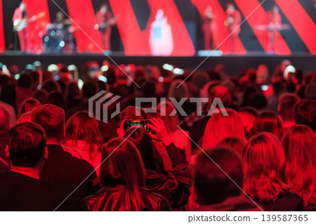 Audience recording live concert with smartphone in red stage lighting, crowd watching band performance Audience recording live concert with smartphone in red stage lighting, crowd watching band performance 139587365