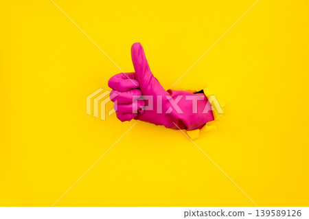 Pink cleaning glove thumbs up through torn yellow paper background Pink cleaning glove thumbs up through torn yellow paper background 139589126
