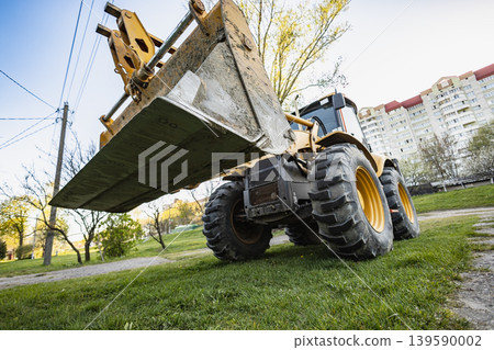 A construction vehicle is lifting dirt in a park setting. Trees are visible around, and buildings are seen in the background under clear skies. Loader 139590002