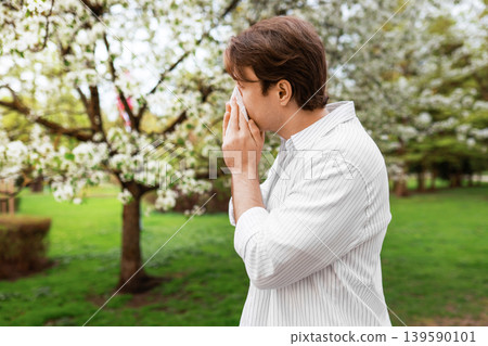 A man sneezes into a tissue while standing near blooming trees in a park. This image depicts seasonal allergies, hay fever, and the impact of spring pollen on health. A man sneezes into a tissue while standing near blooming trees in a park. This image depicts seasonal allergies, hay fever, and the impact of spring pollen on health. 139590101