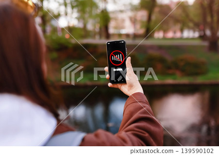 A woman holds a smartphone showing a "No Signal" message while standing in a park near water. The image represents cellular network failure, poor connection, or data loss. A woman holds a smartphone showing a "No Signal" message while standing in a park near water. The image represents cellular network failure, poor connection, or data loss. 139590102