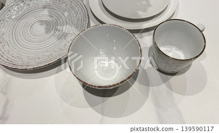 A minimalist set of white and beige ceramic tableware featuring a textured plate, a bowl with a brown rim, and a matching cup on a bright surface A minimalist set of white and beige ceramic tableware featuring a textured plate, a bowl with a brown rim, and a matching cup on a bright surface 139590117