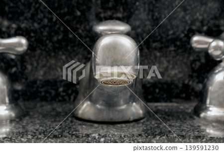 a central silver spout rises with sculptural strength from a backdrop of dark, textured stone. Its rounded form remains in sharp focus, while the flanking cross-handles soften into a gentle blur. This 139591230