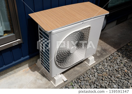 Air conditioner outdoor unit. A wooden board has been placed on top to protect it from the heat. White unit. Air conditioner outdoor unit. A wooden board has been placed on top to protect it from the heat. White unit. 139591347