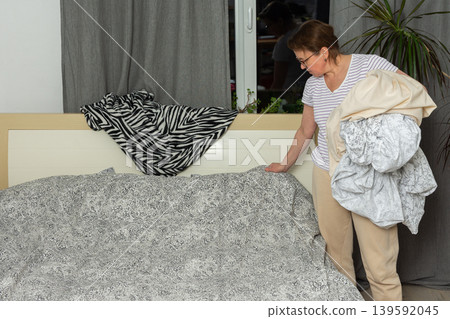 A woman of 53 years old is changing bed linen in her bedroom. She removes old linen and replaces it with new bedding while standing by the bed in daylight A woman of 53 years old is changing bed linen in her bedroom. She removes old linen and replaces it with new bedding while standing by the bed in daylight 139592045