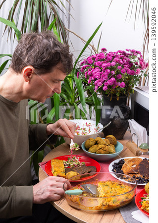 A man sits at a round table and eats various dishes made from homemade recipes. He is surrounded by plants and a bouquet of flowers in a sunny kitchen during breakfast time A man sits at a round table and eats various dishes made from homemade recipes. He is surrounded by plants and a bouquet of flowers in a sunny kitchen during breakfast time 139592046
