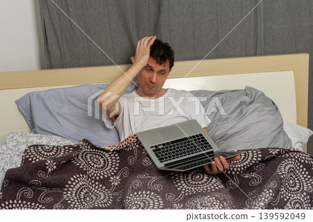 A man stretches while sitting up in bed with a laptop in front of him. It is morning and he has just woken up. The bed appears messy with blankets and sheets around him A man stretches while sitting up in bed with a laptop in front of him. It is morning and he has just woken up. The bed appears messy with blankets and sheets around him 139592049