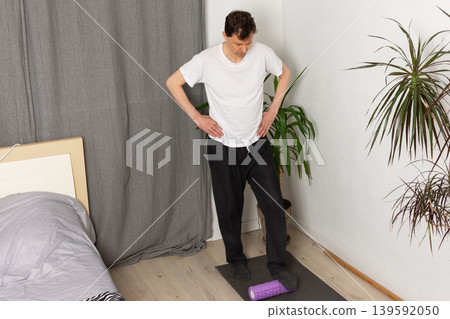 A 53-year-old man is performing exercises at home. He stands on a mat with one foot on a foam roller. There are plants in the background and a simple room setup 139592050