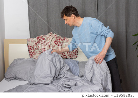 A 53-year-old man is actively changing the bed linen in his bedroom. He is focused on putting fresh sheets on the bed, demonstrating a common home chore 139592053