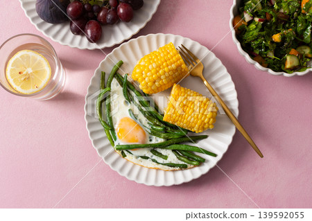 A healthy breakfast is served on a pink table. There are plates with asparagus, a fried egg, corn, and a fresh salad. Grapes and sunflower seeds are on the side A healthy breakfast is served on a pink table. There are plates with asparagus, a fried egg, corn, and a fresh salad. Grapes and sunflower seeds are on the side 139592055