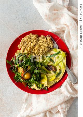 Grains, cooked greens, and vegetables are arranged on a red plate. A bowl of mixed salad with tomatoes is placed nearby on a light surface Grains, cooked greens, and vegetables are arranged on a red plate. A bowl of mixed salad with tomatoes is placed nearby on a light surface 139592063