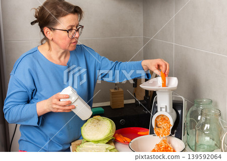 A woman is working in a kitchen to prepare fermented cabbage. She is using a grater and mixing in shredded carrots while measuring salt. The kitchen has different utensils and jars A woman is working in a kitchen to prepare fermented cabbage. She is using a grater and mixing in shredded carrots while measuring salt. The kitchen has different utensils and jars 139592064