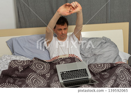 A man stretches while sitting up in bed with a laptop in front of him. It is morning and he has just woken up. The bed appears messy with blankets and sheets around him A man stretches while sitting up in bed with a laptop in front of him. It is morning and he has just woken up. The bed appears messy with blankets and sheets around him 139592065