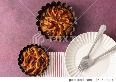 A plum pie is placed on a table with thinly sliced plums visible on top. Bowls of fresh plums are nearby with empty plates waiting for dessert A plum pie is placed on a table with thinly sliced plums visible on top. Bowls of fresh plums are nearby with empty plates waiting for dessert 139592083