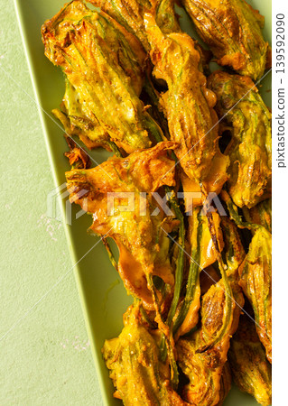 Delicious deep-fried zucchini flowers are neatly arranged on a green platter, showcasing their crispy texture. A bright yellow napkin adds warmth to the scene 139592090