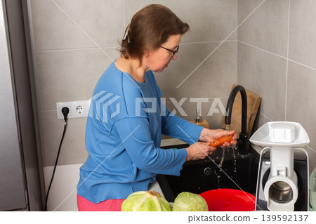 In a bright kitchen, a woman washes carrots at the sink. She focuses on cleaning the vegetables before starting the process of fermenting cabbage In a bright kitchen, a woman washes carrots at the sink. She focuses on cleaning the vegetables before starting the process of fermenting cabbage 139592137