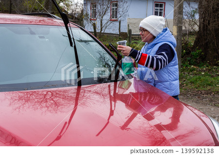 A person is cleaning the windshield of a red car with a cloth while holding a spray bottle. This activity is taking place outside a house on a cloudy day A person is cleaning the windshield of a red car with a cloth while holding a spray bottle. This activity is taking place outside a house on a cloudy day 139592138