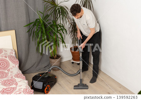 A 53-year-old man uses a vacuum cleaner to clean the floor in a room decorated with houseplants. He focuses on keeping the space tidy and organized while working 139592263