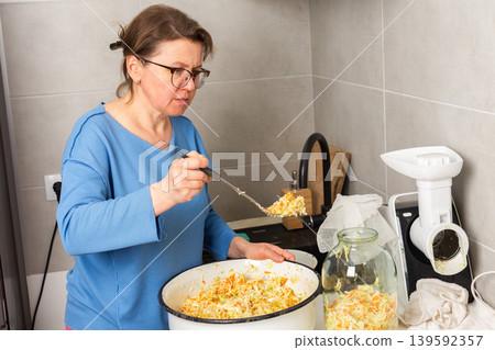 A woman stands in her kitchen, mixing cabbage for fermentation. She uses a spoon to stir the cabbage while readying jars for storage. The atmosphere is filled with food preparation A woman stands in her kitchen, mixing cabbage for fermentation. She uses a spoon to stir the cabbage while readying jars for storage. The atmosphere is filled with food preparation 139592357