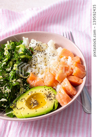 Fresh breakfast bowl featuring salmon, sliced avocado, leafy greens, and rice served on a pink striped tablecloth. Perfect for a nutritious start to the day Fresh breakfast bowl featuring salmon, sliced avocado, leafy greens, and rice served on a pink striped tablecloth. Perfect for a nutritious start to the day 139592435