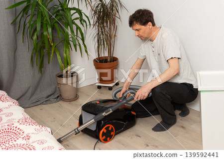 A 53-year-old man is getting ready to clean the floor with a vacuum cleaner. He is kneeling in a room that has plants and a bed nearby. The area looks tidy and organized A 53-year-old man is getting ready to clean the floor with a vacuum cleaner. He is kneeling in a room that has plants and a bed nearby. The area looks tidy and organized 139592441