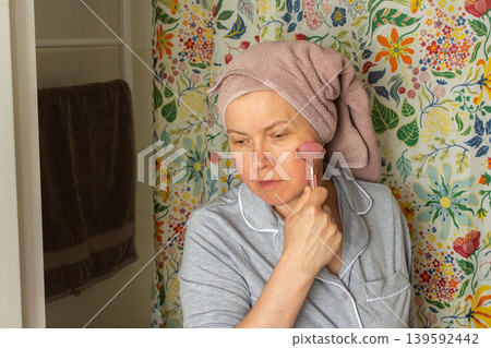 A 50 year old woman applies a facial roller to her face while standing in a bright bathroom. She has a towel wrapped around her hair and wears comfortable clothes A 50 year old woman applies a facial roller to her face while standing in a bright bathroom. She has a towel wrapped around her hair and wears comfortable clothes 139592442